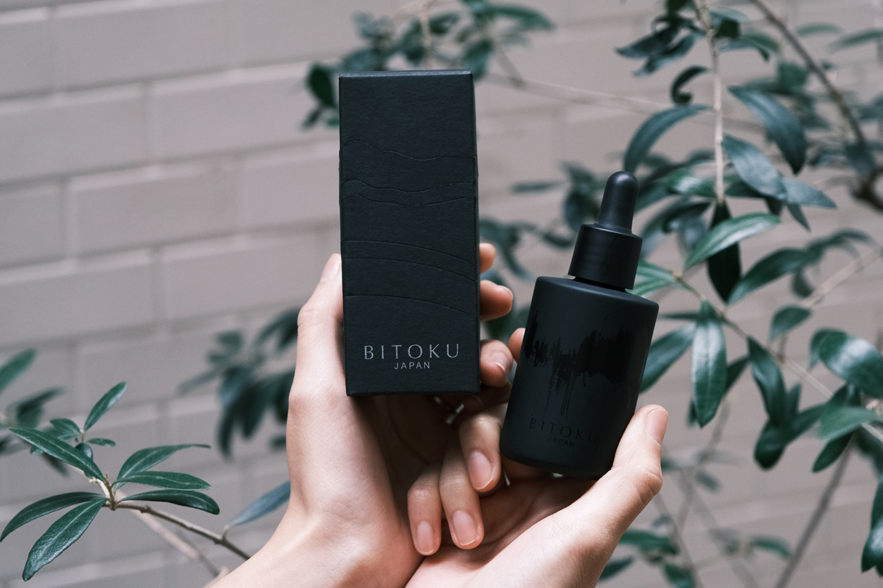 植物を背景に、両手で持ったBITOKUの黒い化粧箱と美容液ボトル